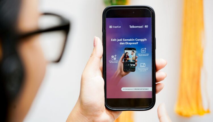 Seorang pengguna yang sedang menggenggam ponsel pintar menampilkan visual resmi peluncuran kerja sama bertuliskan "CapCut | Telkomsel 5G - Edit jadi Semakin Canggih dan Ekspresif" lengkap dengan penjelasan fitur bertenaga AI.